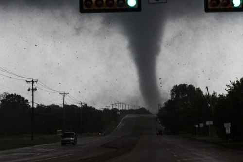A tornado forms in the distance over a road, with dark clouds and a green traffic light visible.