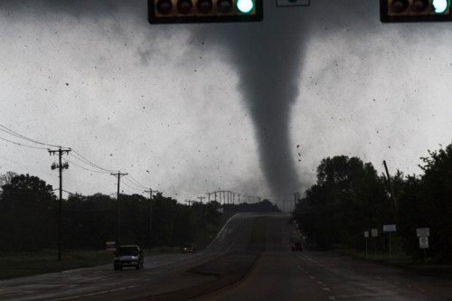 A tornado forms in the distance over a road, with dark clouds and a green traffic light visible.