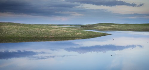A serene landscape featuring a calm river reflecting the sky, surrounded by green hills and grassy banks.