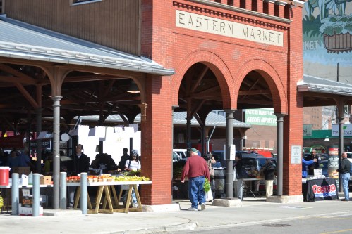 A bustling market scene at Eastern Market, featuring vendors, shoppers, and colorful produce under a brick archway.