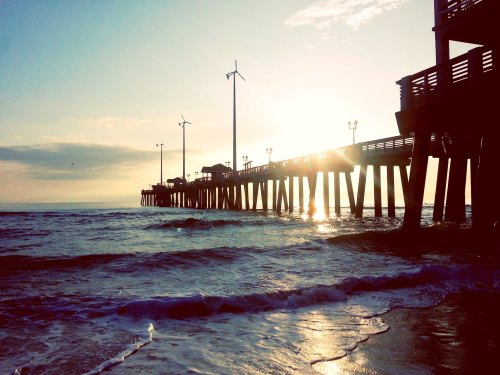 Silhouette of a pier at sunset, with wind turbines and gentle waves lapping at the shore.