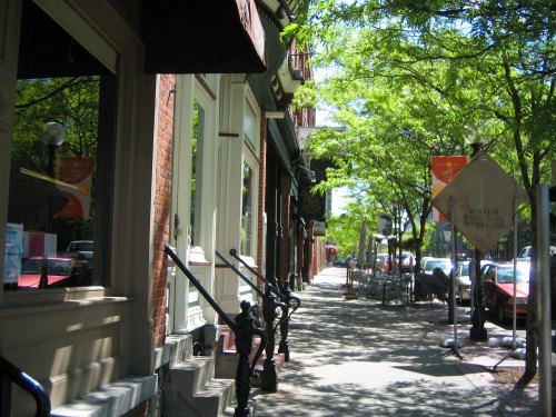 A sunny street lined with brick buildings, green trees, and sidewalk cafes, creating a vibrant urban atmosphere.
