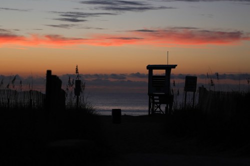Silhouette of a lifeguard tower at sunset, with colorful clouds over the ocean and beach grass in the foreground.