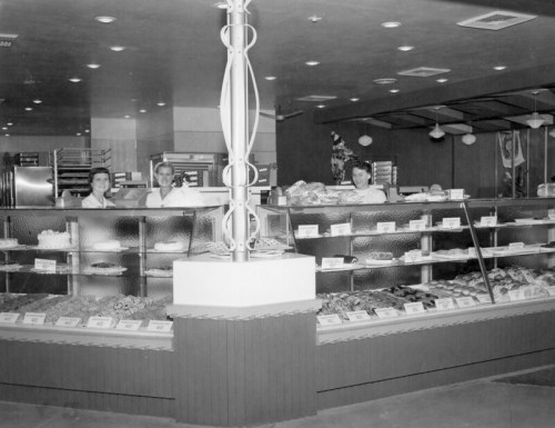 Black and white photo of a bakery with three staff members behind a display of various baked goods.