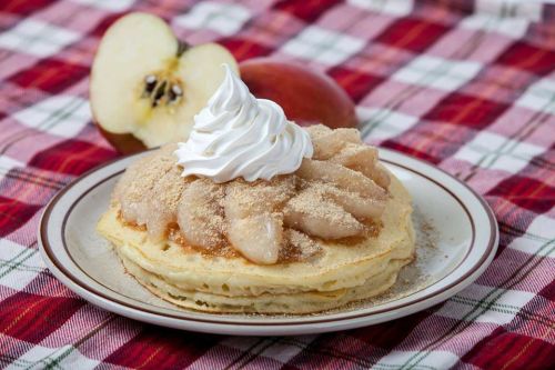 A plate of pancakes topped with apple slices, cinnamon, and whipped cream, with a fresh apple in the background.