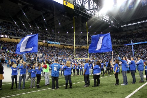 A crowd of fans in blue shirts wave large flags at a sports stadium during a game, with cheering spectators in the background.