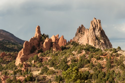 Rocky formations rise amidst green trees under a cloudy sky, showcasing a dramatic natural landscape.