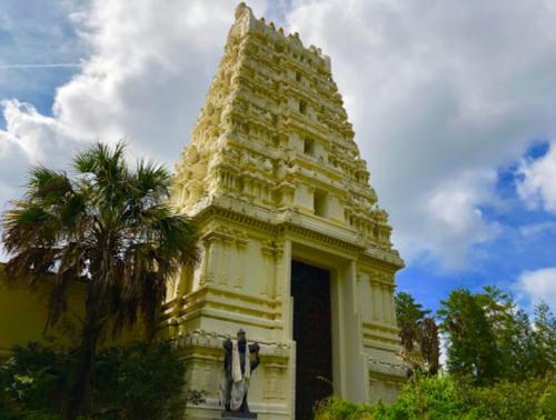 A tall, ornate temple tower with intricate carvings, surrounded by greenery and under a partly cloudy sky.