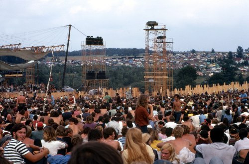 A large crowd at an outdoor music festival, with stages and a distant view of a town in the background.