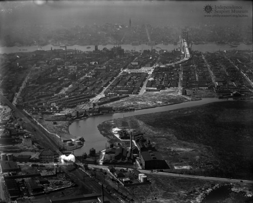 Aerial view of a cityscape with industrial buildings, a river, and a bridge in the distance, captured in black and white.