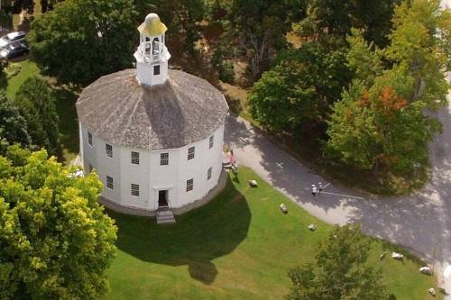 Aerial view of a round white building with a bell tower, surrounded by trees and a grassy area.