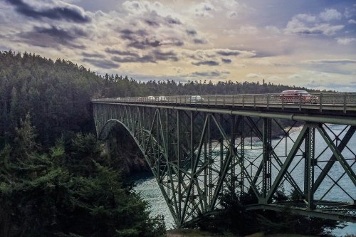 A green metal bridge spans a river, surrounded by trees and under a cloudy sky. Vehicles are visible on the bridge.