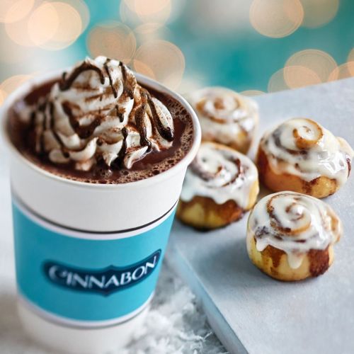 A cup of hot chocolate topped with whipped cream, alongside several small cinnamon rolls on a light surface.