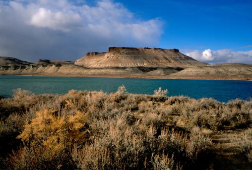 A scenic view of a plateau rising above a turquoise lake, surrounded by dry shrubs and a blue sky with clouds.