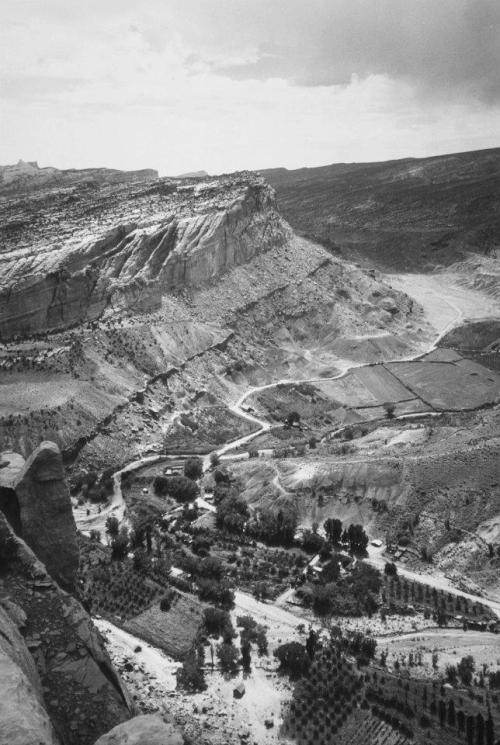 A black and white landscape of rugged cliffs and a winding river, surrounded by fields and trees in a valley.