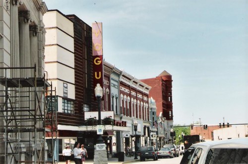 A street view featuring historic buildings, shops, and a sign for "WOGU" under a clear blue sky.