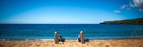 Two scuba tanks and gear sit on sandy beach, with calm blue ocean and clear sky in the background.
