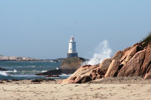 A lighthouse stands on a rocky shore, with waves crashing against the rocks and a clear blue sky above.
