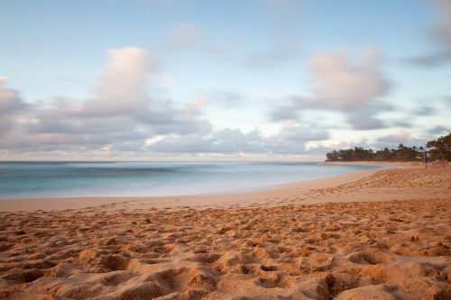 A serene beach scene with soft sand, gentle waves, and a cloudy sky in the background.