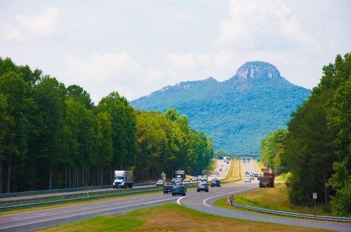 A highway lined with trees, featuring cars and trucks, with a mountain in the background under a blue sky.