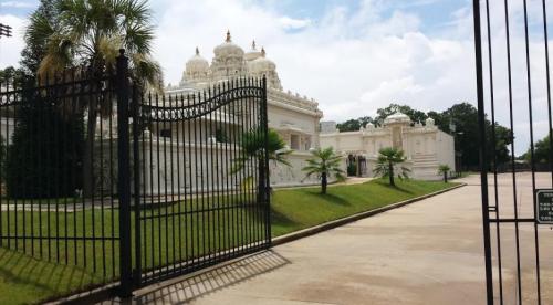A white temple with ornate domes, surrounded by greenery and a black gate, under a partly cloudy sky.