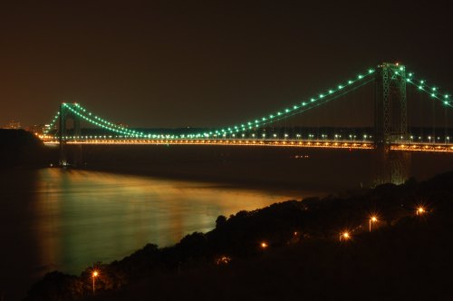 A brightly lit suspension bridge at night, reflecting in calm waters below, surrounded by dark trees and city lights.