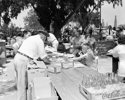 A black-and-white photo of people at tables, engaging in activities outdoors, with trees and oil rigs in the background.