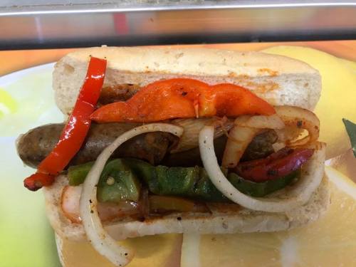A sausage in a bun topped with grilled onions and colorful bell peppers.