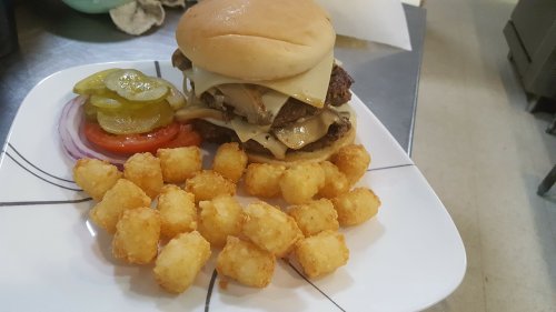 A juicy burger with melted cheese, pickles, tomato, and onion, served with crispy tater tots on a plate.