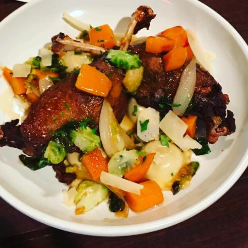 A bowl of roasted duck with vegetables, including carrots, Brussels sprouts, and onions, garnished with herbs.