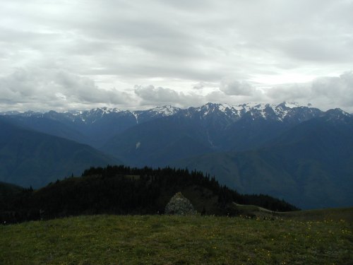 A panoramic view of rugged mountains under a cloudy sky, with green hills and patches of snow visible on the peaks.