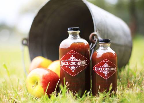 Two bottles of Athol Orchards cider syrup sit on grass near fresh apples, with a metal bucket in the background.