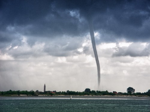 A dark tornado descends from stormy clouds over a calm body of water, with a distant shoreline and a tower visible.