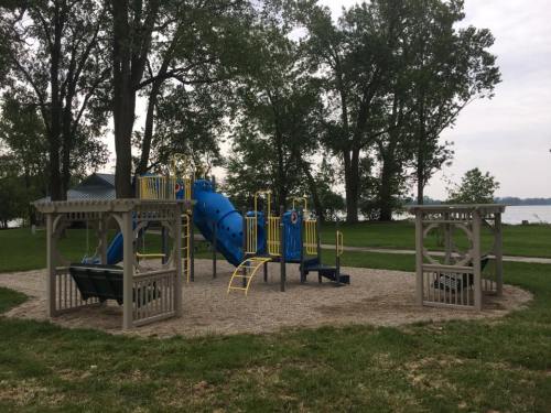 A playground with a blue slide and climbing structures, surrounded by trees and near a lake.