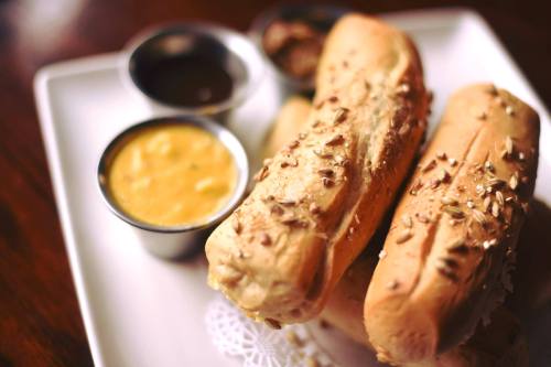 Three freshly baked bread rolls topped with seeds, served with small bowls of mustard and sauce on a white plate.