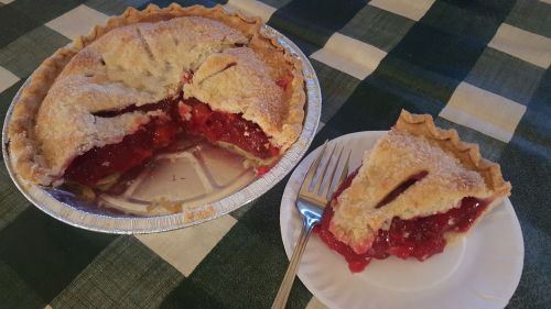 A cherry pie with a slice removed, sitting on a plate next to the pie dish, on a green and blue checkered tablecloth.