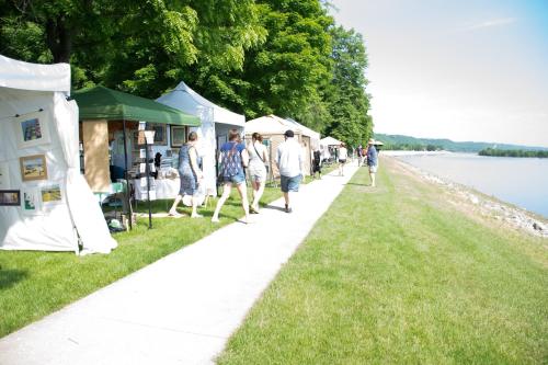 People stroll along a riverside path lined with tents at an outdoor market or festival. Green trees in the background.