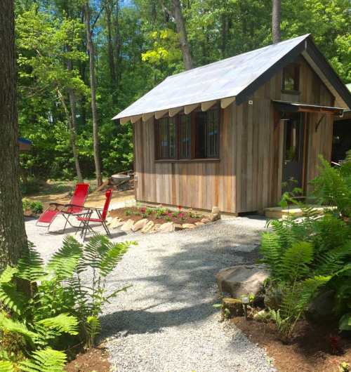 A small wooden cabin with a metal roof, surrounded by greenery and gravel, featuring two red chairs in front.