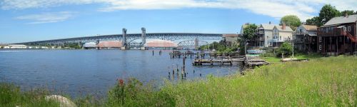 A panoramic view of a river with a bridge, grassy shore, and houses along the water's edge under a blue sky.