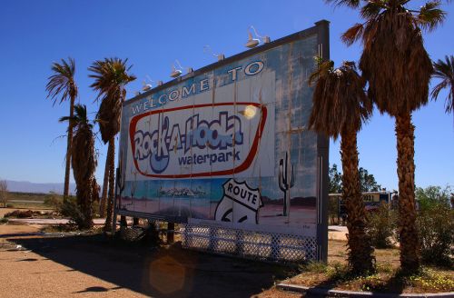 A weathered sign welcomes visitors to Rock-A-Hoola Waterpark, surrounded by palm trees and a clear blue sky.