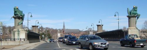 A busy bridge with frog sculptures on pillars, surrounded by cars and a church in the background under a clear blue sky.