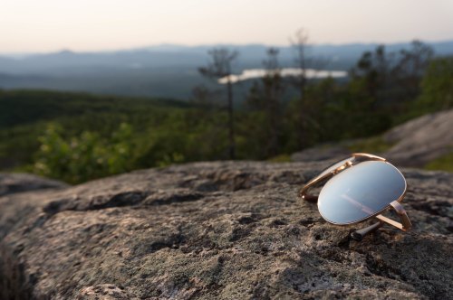 Sunglasses resting on a rock, with a scenic view of mountains and a lake in the background during sunset.