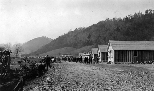 Historic black-and-white photo of a dirt road lined with wooden buildings and people walking, set against a mountainous backdrop.