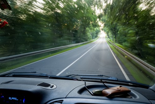 A blurred view of a winding road through trees, seen from the dashboard of a vehicle. Sunglasses rest on the dashboard.