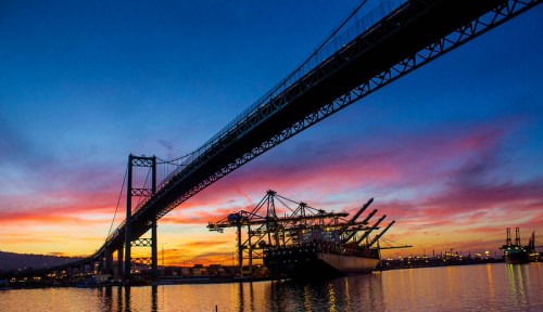Silhouette of a bridge over a harbor at sunset, with cargo ships and cranes against a colorful sky.