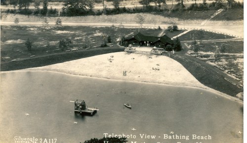 Aerial view of a bathing beach with a sandy area, a building, and boats on the water. Trees in the background.