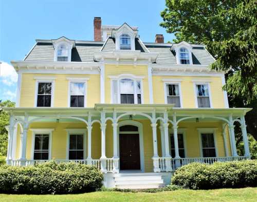 A large yellow Victorian house with a white porch and green trees in the background under a clear blue sky.