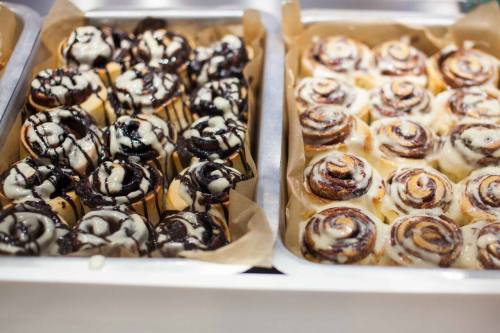 Two trays of cinnamon rolls: one with chocolate drizzle and the other with cream cheese icing.