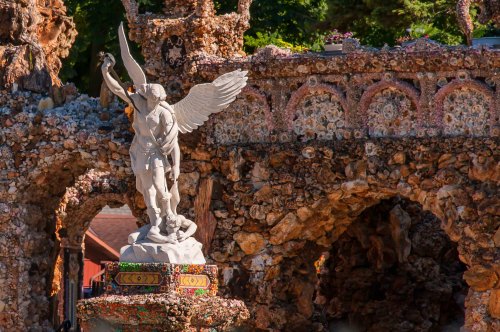 A stone statue of an angel with wings stands atop a colorful, ornate base amidst a rocky, textured backdrop.