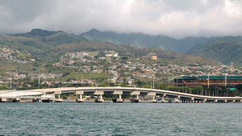 A bridge spans a body of water, with lush green mountains and residential areas in the background under a cloudy sky.